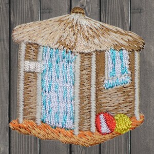 Peut inclure: Patch brodé représentant une cabane de plage brune avec un toit de chaume, des rideaux rayés bleus et blancs, et un ballon de plage rouge et blanc et un ballon de plage jaune devant.