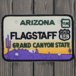Op de afbeelding: Witte en gele geborduurde patch met de tekst "Arizona", "Flagstaff", "Grand Canyon State" en "Route 66". De patch toont een paarse bergketen met een cactus en een saguaro-cactus.