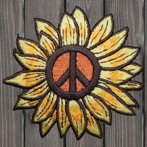 Puede incluir: Un girasol amarillo con un símbolo de paz marrón en el centro. El girasol tiene un contorno marrón oscuro y está bordado.