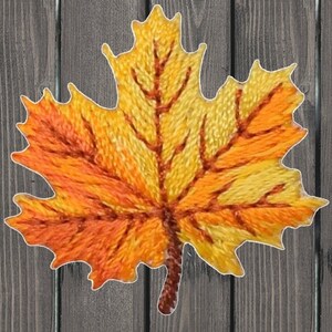 以下が含まれることがあります： オレンジ、イエロー、ブラウンのステッチで刺繍された秋の葉のクローズアップ。葉は、詳細な葉脈パターンを持つメープルリーフです。