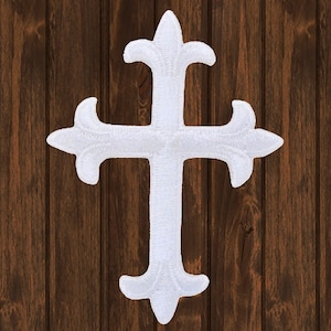 Puede incluir: Parche de cruz bordado en blanco con un diseño decorativo. La cruz está sobre un fondo de madera marrón.