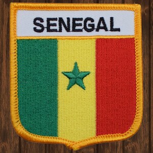 May include: A yellow, green, and red embroidered patch with a gold border. The patch features a green star on a yellow background and the word "SENEGAL" in black text on a white background.