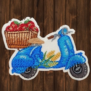 May include: Embroidered patch featuring a blue scooter carrying a basket of red strawberries. The scooter has wheat stalks and a brown handlebar. The patch is on a white background with a brown wooden backdrop.