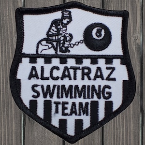 Könnte beinhalten: Schildförmiger Aufnäher mit schwarzem Rand und weißem Hintergrund. Der Aufnäher zeigt einen Gefangenen, der an einer 8-Ball-Kugel angekettet ist, mit den Worten "ALCATRAZ SWIMMING TEAM" in schwarzen Buchstaben.