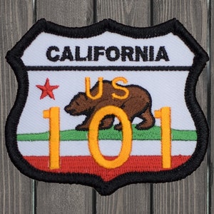 May include: A white, red, and green embroidered patch with a black border. The patch is shaped like a shield and features the text "CALIFORNIA" at the top, a red star, a brown bear, and the text "US 101" in yellow.