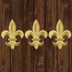 May include: Three gold fleur-de-lis designs on a dark brown wood background.