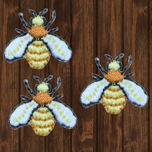 Puede incluir: Tres parches bordados de abejas con rayas amarillas y marrones, alas blancas y antenas negras. Las abejas están sobre un fondo de madera marrón.