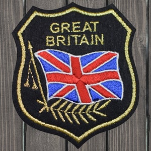 May include: A black felt patch with gold stitching in the shape of a shield. The patch features a gold embroidered Union Jack flag with the words "GREAT BRITAIN" above it. The flag is on a gold pole with a gold tassel. There are gold embroidered leaves at the bottom of the patch.