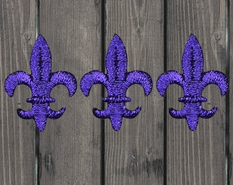 May include: Three purple embroidered fleur-de-lis patches on a wood background.