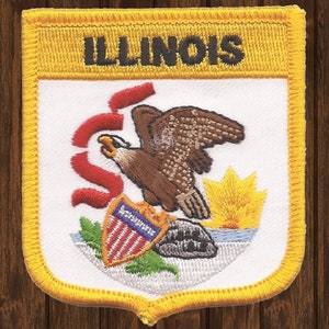 May include: A white embroidered patch with a yellow border. The patch features the state seal of Illinois, which includes an eagle, a shield with stars and stripes, a sunrise, and a red ribbon. The text "ILLINOIS" is embroidered in black on a yellow banner at the top of the patch.