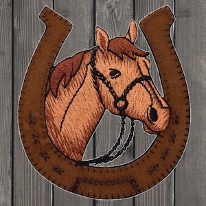 May include: Embroidered patch featuring a horse head within a horseshoe. The horse is brown with a black bridle, set against a brown horseshoe. The background is a rustic wooden texture, creating a Western or equestrian theme.