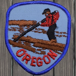 Op de afbeelding: Een blauwe en rode geborduurde patch met een man in een rood en zwart shirt die een lange stok vasthoudt, staand op een boomstam in een rivier. De tekst "OREGON" staat in rode letters op de patch.