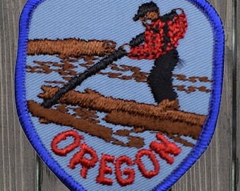 May include: A blue and red embroidered patch with a man in a red and black shirt holding a long pole, standing on a log in a river. The text "OREGON" is in red letters on the patch.