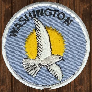 May include: A white embroidered patch with a blue background and white stitching. The patch features a white dove flying in front of a yellow sun with the word "WASHINGTON" in black stitching around the edge.