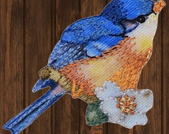 Può includere: Un passero azzurro ricamato con piume arancioni e gialle, appollaiato su una foglia verde con un fiore bianco. L'uccello ha un occhio nero e un becco bianco.