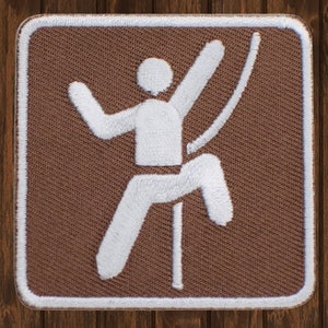 May include: A brown and white embroidered patch featuring a stylized figure climbing a rope. The patch has a square shape with rounded corners and a white border. The climber is white against the brown background.