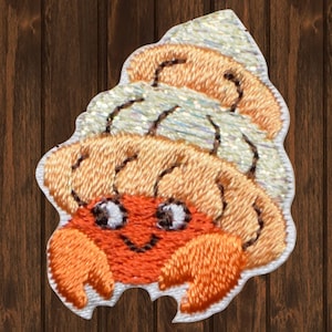 May include: A small, orange and brown embroidered patch of a hermit crab with a sparkly shell. The crab is smiling and has large, white eyes.