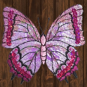 May include: A purple and pink butterfly with iridescent sequins, embroidered with black lines and pink accents. The butterfly is on a brown wooden background.