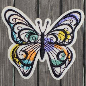May include: Embroidered butterfly patch with a cream-colored background. The butterfly features black outlines and colorful details in shades of blue, orange, yellow, and purple. The patch is designed with intricate patterns.