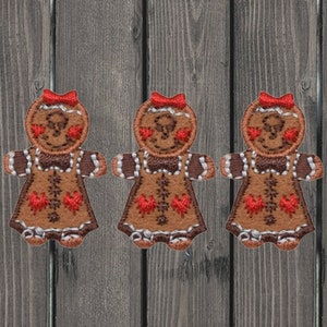May include: Three gingerbread person appliques with red bows, white trim, and red hearts on their faces. The appliques are brown and have a white and red pattern on their dresses.
