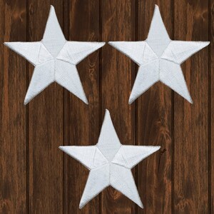 Puede incluir: Tres parches blancos en forma de estrella con costuras detalladas. Las estrellas están dispuestas sobre un fondo de madera marrón oscuro. Estos parches se pueden utilizar con fines decorativos o para embellecer ropa y accesorios.