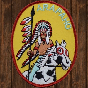 Könnte beinhalten: Ein gelber gestickter Aufnäher mit rotem Rand. Der Aufnäher zeigt einen Indianer, der auf einem Pferd reitet. Der Mann trägt einen Federkopfschmuck und hält eine Lanze. Der Text "ARAPAHO" ist in blauer Schrift um den Rand des Aufnähers geschrieben.