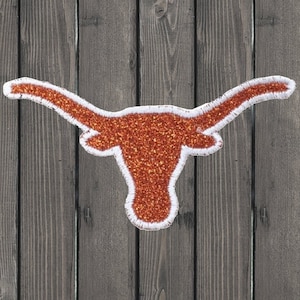 May include: A large, orange, glitter-covered, felt applique of a longhorn steer head with white outlines. The applique is on a wood background.