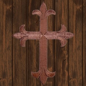 Pode incluir: Uma cruz marrom ornamentada com um design de flor-de-lis em cada extremidade. A cruz está contra um fundo de grão de madeira.