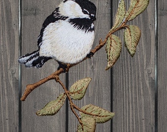 May include: Embroidered black and white chickadee perched on a branch with green leaves. The bird is sitting on a branch with green leaves.