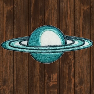 May include: An embroidered patch of the planet Saturn, featuring a teal blue colour and white rings. The patch is displayed against a brown wooden backdrop.