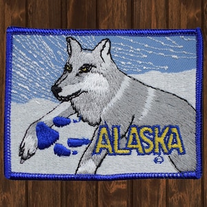 Könnte beinhalten: Gestickter Aufnäher mit einem weißen Wolf mit blauen Pfotenabdrücken und dem Wort "ALASKA" in gelben Buchstaben auf blauem Hintergrund.