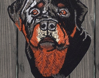 May include: Embroidered patch of a Rottweiler dog's head. The dog has black fur with brown markings around the eyes and mouth. The patch is made of black fabric with detailed embroidery.