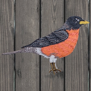 May include: Embroidered patch of a red-breasted robin with a black head and yellow beak, perched on a wooden background.