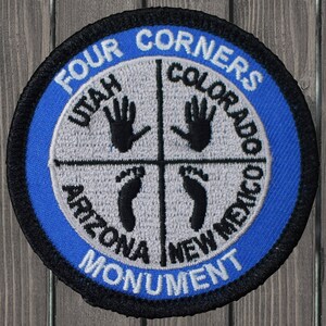 May include: A blue and black embroidered patch with the text "Four Corners Monument" and the names of the four states that meet at the monument: Utah, Colorado, Arizona, and New Mexico. Each state is represented by a handprint or footprint.
