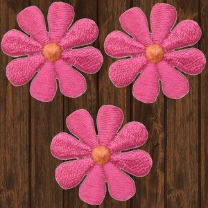 May include: Three pink fabric flower appliques with a yellow centre. The flowers are on a brown wooden background.