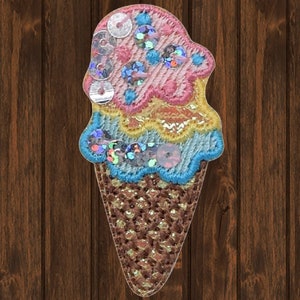 May include: A colorful embroidered ice cream cone patch with pink, blue, and yellow layers of ice cream decorated with sequins. The cone is brown and has a textured pattern.