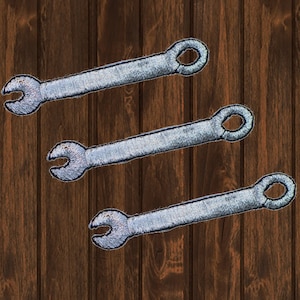 Puede incluir: Tres parches termoadhesivos en forma de llave inglesa plateada. Los parches están dispuestos en una pila vertical sobre un fondo de madera marrón.