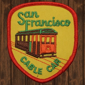 Puede incluir: Un parche bordado amarillo con borde rojo. El parche presenta un tranvía con el texto "San Francisco Cable Car" en verde.