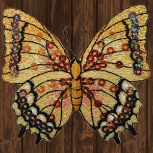 May include: A decorative butterfly applique in golden yellow, with wings embellished with orange, red, and iridescent sequins, and outlined in black. The butterfly is set against a dark brown wooden background.