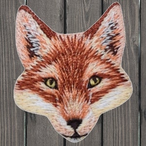 May include: Embroidered fox head patch with a detailed design. The fox has a reddish-brown fur with white accents around the muzzle and ears. The eyes are yellow and the nose is black.