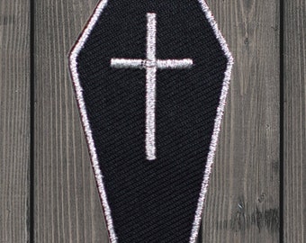 Pode incluir: Patch de tecido preto em forma de caixão com uma cruz branca bordada. O patch tem um contorno branco.