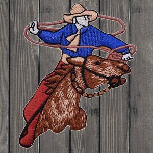 Puede incluir: Parche bordado de un vaquero montando un caballo marrón, con una camisa azul, pantalones rojos y un sombrero marrón. El vaquero sostiene un lazo en su mano derecha.
