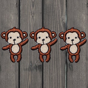 以下が含まれることがあります： 白い顔と茶色の腕と脚を持つ、茶色の刺繍された猿のアップリケが3つ。猿は腕を伸ばして立っています。