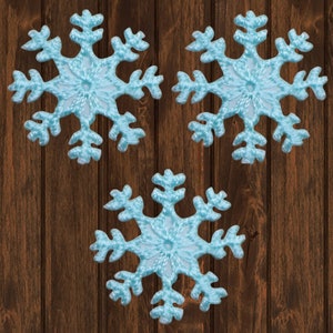 Puede incluir: Tres adornos de copo de nieve azul claro sobre un fondo de madera marrón.