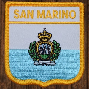 Op de afbeelding: Een gele geborduurde patch met een witte achtergrond en een blauwe en witte vlag van San Marino. De vlag heeft een wapenschild met een kroon, drie torens en een lauwerkrans. De tekst "SAN MARINO" is in het geel geborduurd bovenaan de patch.