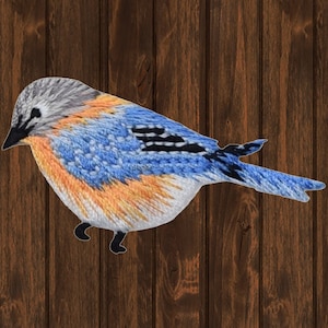 May include: Embroidered patch of a bluebird with gray, blue, and orange plumage. The bird is depicted in a side profile, with its wings and tail spread. The patch is set against a wooden background.