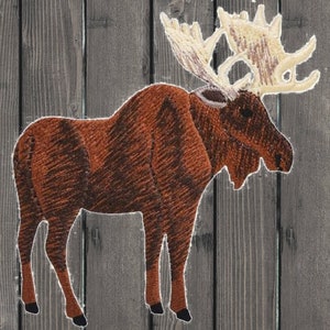 Op de afbeelding: Een geborduurde bruine eland met groot gewei, tegen een grijze houten achtergrond. De eland heeft een gedetailleerde textuur en het gewei is lichtbeige van kleur. De afbeelding is een close-up van de eland.