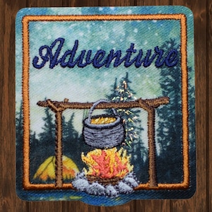 May include: Embroidered patch with a blue sky and forest background. The text "Adventure" is stitched in blue thread. A campfire with a pot hanging over it is in the center of the patch.