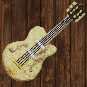 Può includere: Toppa ricamata di una chitarra elettrica gialla con corde nere e paletta marrone. La chitarra presenta un design dettagliato con un corpo color crema e dettagli marroni. La toppa è posta su uno sfondo di venature del legno scuro.
