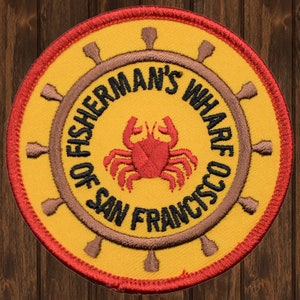 May include: A yellow and red embroidered patch with a brown wheel and the text "Fisherman's Wharf of San Francisco". A red crab is in the center of the patch.
